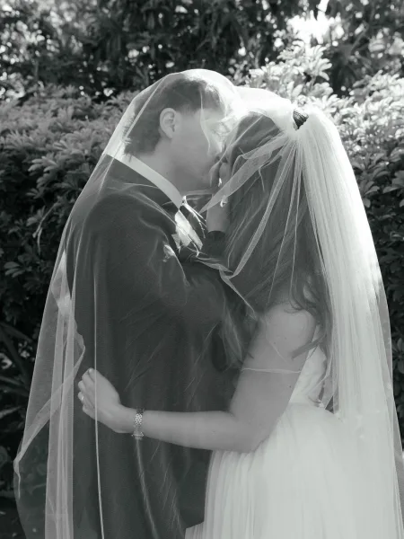 Wedding kiss as bride and groom kiss under a bridal veil, his dark suit and boutonniere framed by lush garden greenery