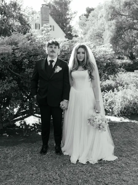 Couple portrait, black and white wedding portrait of bride and groom holding hands on a lawn, her veil trailing beside garden shrubs and a house