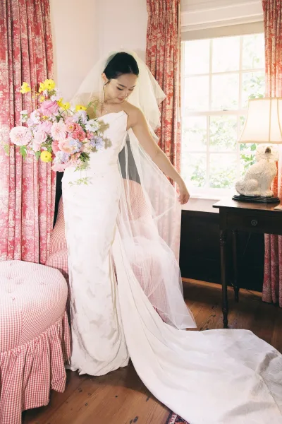 Bridal portrait of bride holding bouquet in a strapless wedding dress, long veil and train, looking down by a window with patterned curtains