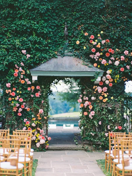 Ceremony entrance decor with a floral arch entrance of garden roses and climbing ivy framing a stone archway and aisle with chairs on lawn