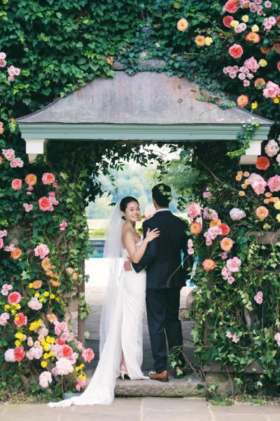Couple portrait of bride and groom embrace under an ivy-covered floral arch, bride looking back in veil on garden walkway by pool