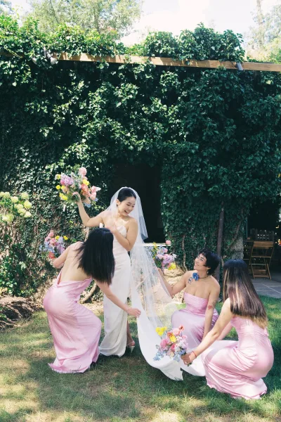 Bridesmaid moment as bridesmaids help the bride with bridesmaids fixing veil, holding colorful bouquets by an ivy wall outdoors