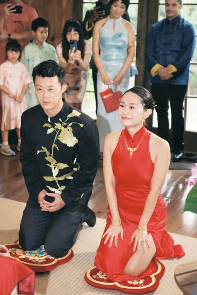 Ceremony moment as couple kneels for a tea ceremony, bride in red cheongsam with gold jewelry, guests filming in wooden room with windows