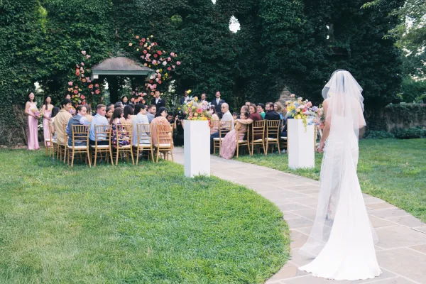 Wedding processional with bride walking down aisle in a cathedral veil past floral arrangements toward groom in tux under a garden gazebo.