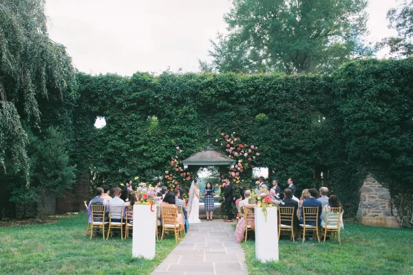 Outdoor wedding ceremony with bride and groom under a floral arch, gold chiavari chairs lining an aisle runner by an ivy-covered wall