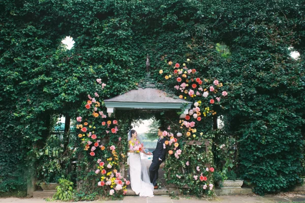 Wedding ceremony moment as bride and groom hold hands at the altar under a round rose floral arch, with veil and ivy wall backdrop
