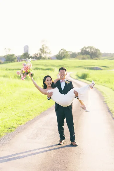 Couple portrait of groom carrying bride as she raises a pink bouquet, laughing on a country road with green fields and a barn silo behind