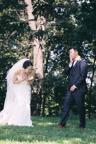 First look moment outdoors as the groom in a navy suit reacts while the bride laughs in a strapless dress with veil in sunlight
