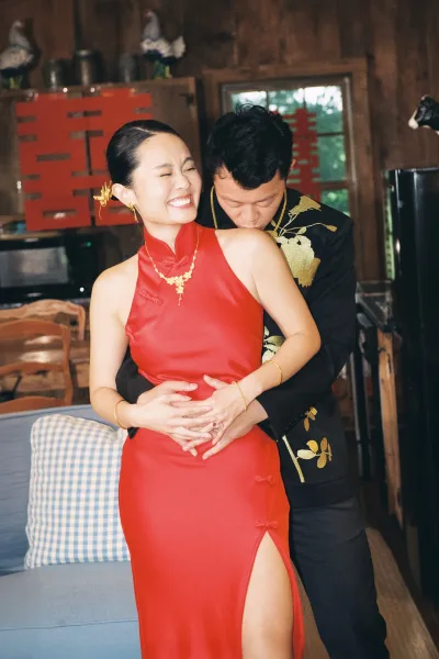 Couple portrait of bride and groom embrace, she in red cheongsam with gold jewelry, him in black embroidered suit inside a wood cabin