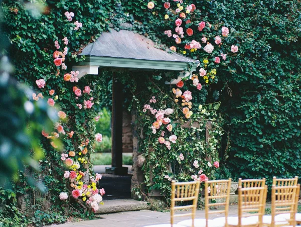 Ceremony backdrop with a floral garland of climbing roses and ivy, mixed pink and yellow blooms on stone steps beside wooden chairs in a garden aisle