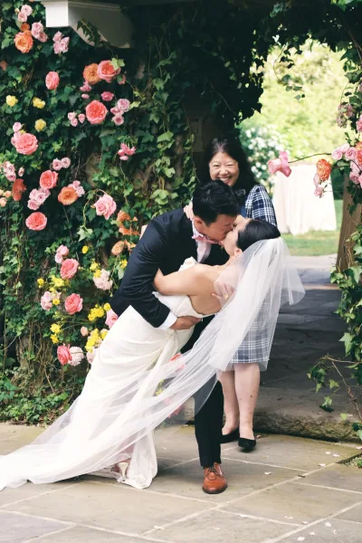 Wedding kiss portrait of groom dipping bride as her long veil blows, framed by a rose and greenery floral arch on a stone garden walkway