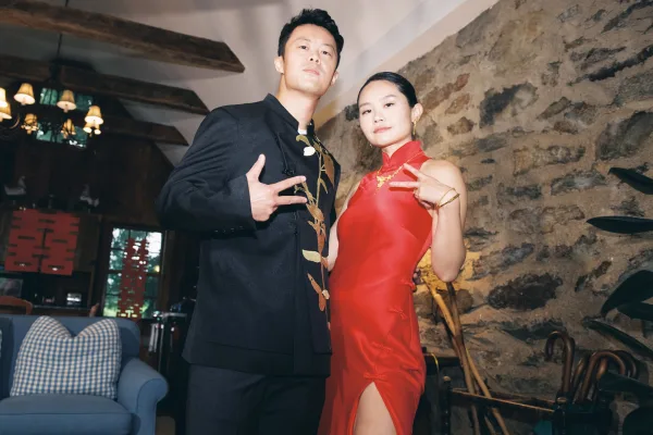 Couple portrait in a Chinese wedding portrait style, bride in red cheongsam with gold jewelry beside groom in black embroidered suit by stone wall