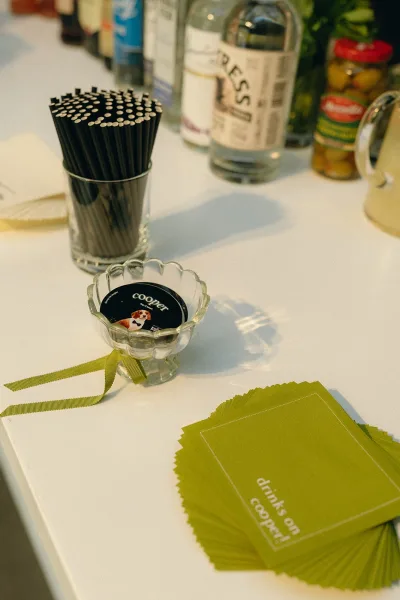 Wedding cocktail napkins with personalized bar details, flanked by cocktail stirrers on a bar counter with liquor bottles and fresh herbs.