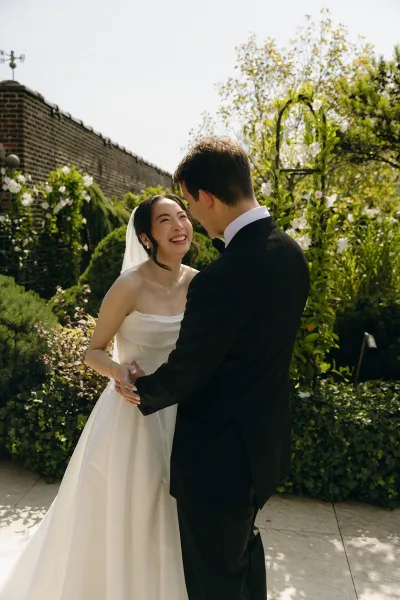 Couple portrait at a wedding first look, bride in a strapless gown with a classic veil meets her groom amid flowering garden vines.