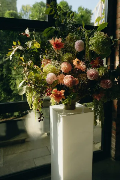Wedding flower arrangement, a pedestal flower arrangement with lush, asymmetrical form, set by a window and brick wall overlooking garden trees.