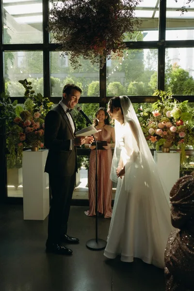 Wedding vows with the groom reading vows to his smiling bride, a vow book in hand, framed by glass windows and garden greenery.
