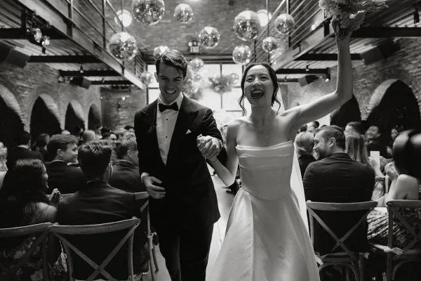 Wedding recessional captures a joyful ceremony exit as newlyweds walk hand in hand, bride lifting a bouquet, amid brick walls and arched alcoves.
