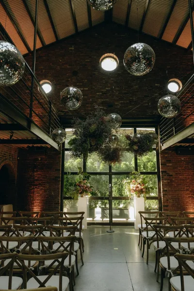Ceremony setup with indoor ceremony decor and a disco ball installation against brick walls and large windows beneath a vaulted ceiling.