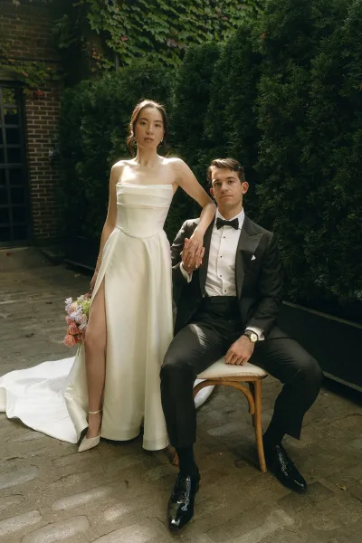 Couple portrait with the bride holding bouquet and the groom at her side, posed against an ivy-clad brick wall and evergreen hedge.