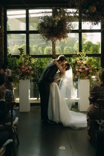 Ceremony kiss marks their first kiss as newlyweds, groom dipping the bride beneath a disco ball, framed by large windows and garden greenery.