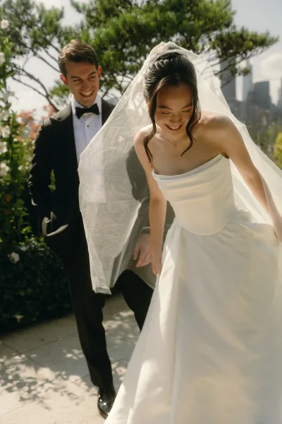 Couple portrait of a bride and groom laughing, her veil catching light in a garden with a distant city skyline behind them.