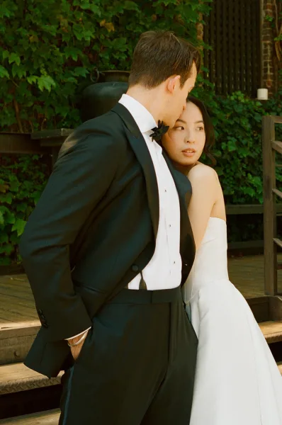 Couple portrait of a bride embracing her groom by an ivy wall and wooden steps, highlighting his black bow tie.
