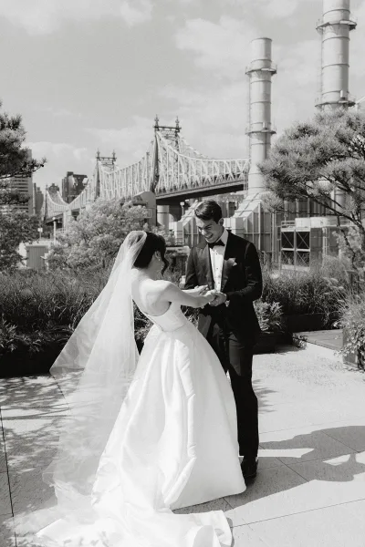 Couple portrait of a bride and groom holding hands on a terrace by a bridge and city buildings, her veil drifting in the breeze.