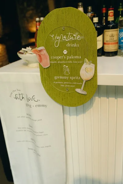 Signature cocktail sign for the wedding bar menu with a paw print icon, set on a bar counter with liquor bottles against a dark backdrop.