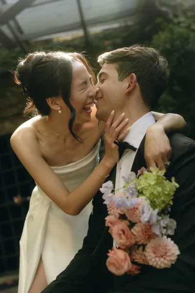 Wedding kiss portrait capturing a tender couple portrait as they lean nose to nose, the bride holding a pastel bouquet against an ivy wall.