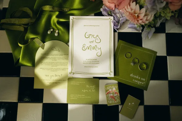 Invitation suite featuring olive green wedding invitations, accented by wedding rings on checkered tile.