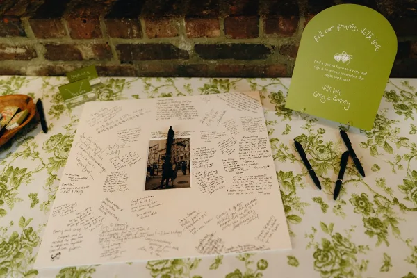 Guest book alternative with a photo mat guest book ready for signatures, set with pens on a styled table against a warm brick wall backdrop.