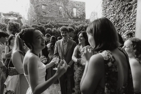 Wedding cocktail hour guests mingle at an outdoor cocktail hour beneath string lights in a courtyard by an ivy wall.