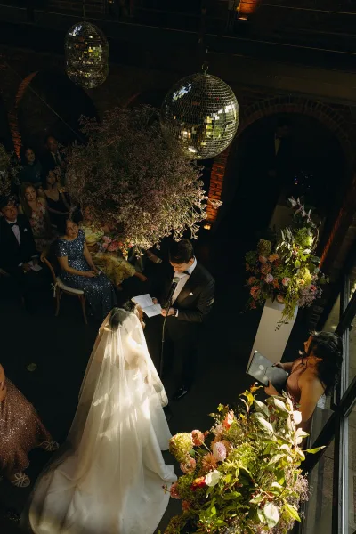 Ceremony moment as a couple exchanging vows beneath brick arches and tall windows, framed by disco balls.