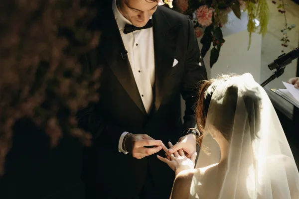 Wedding ring exchange as the groom places the ring on the bride’s finger, her bridal veil floating before a floral backdrop in an indoor venue.