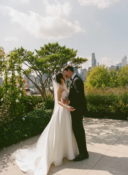 Wedding kiss portrait as a bride and groom kiss, her long veil draped, on a stone patio framed by garden trees and a distant city skyline.