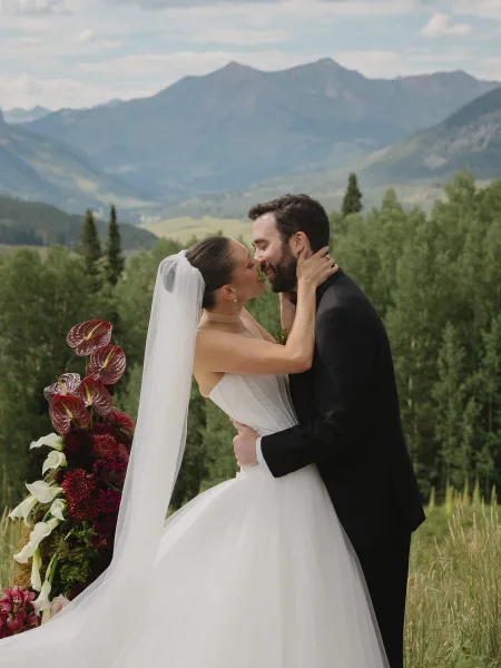 Wedding kiss portrait captures a ceremony kiss moment as the couple embrace, her veil trailing, against a mountain range and evergreens.