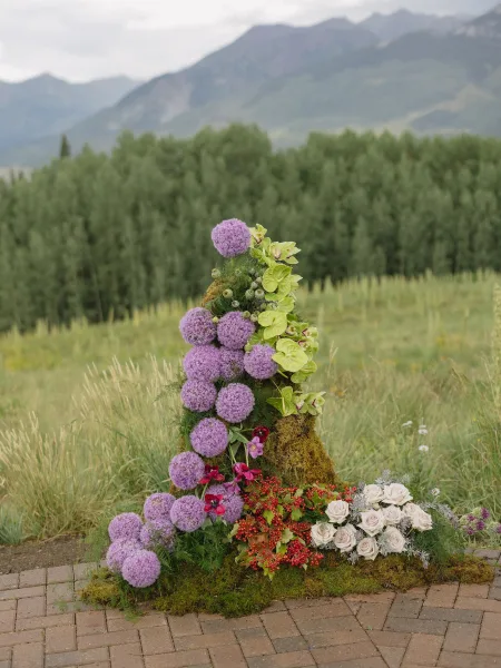 Ceremony floral installation featuring grounded altar flowers with purple allium, set on brick pavers before a mountain range and evergreens.