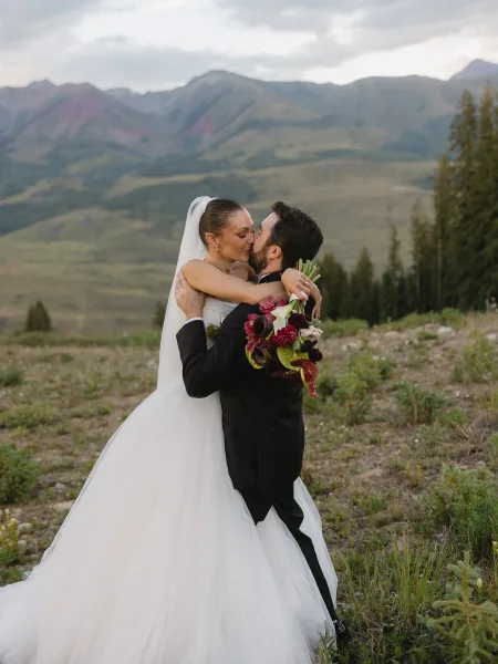 Wedding kiss portrait as the groom lifts the bride in a pine-lined mountain meadow under a cloudy sky, her long veil trailing behind them.