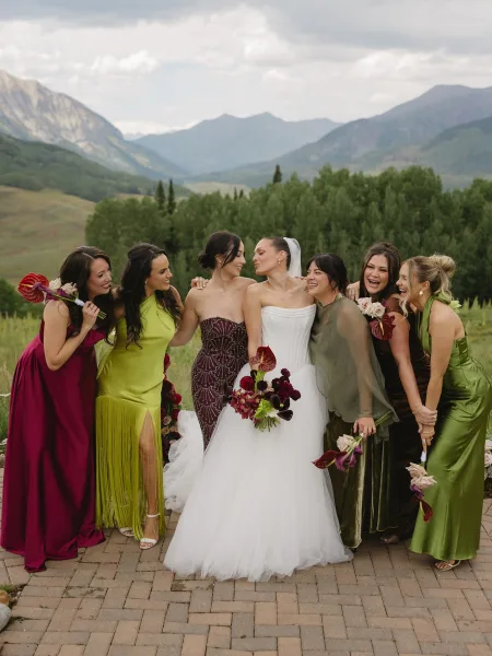 Bride and bridesmaids in a mix-and-match look laugh as they hold calla lily bouquets before evergreen trees and mountains under clouds.