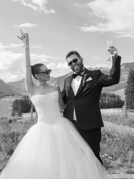 Wedding champagne toast in a black and white wedding photo as the couple raises a toast in sunglasses against a mountain backdrop in a grassy field.