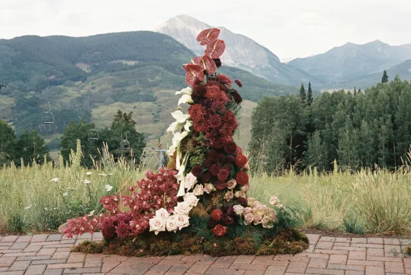 Ceremony floral installation creating a mountain ceremony backdrop, anchored with sculptural blooms and dahlias amid meadow grass and evergreens.