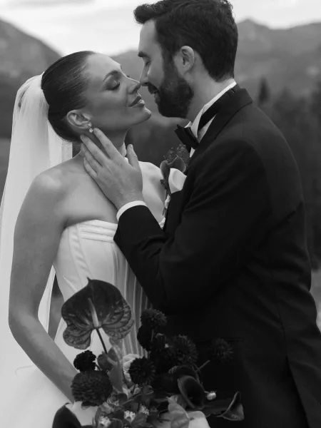 Wedding kiss portrait of a bride and groom close up, leaning in beneath the bride's veil, framed by distant mountains and trees under open sky.