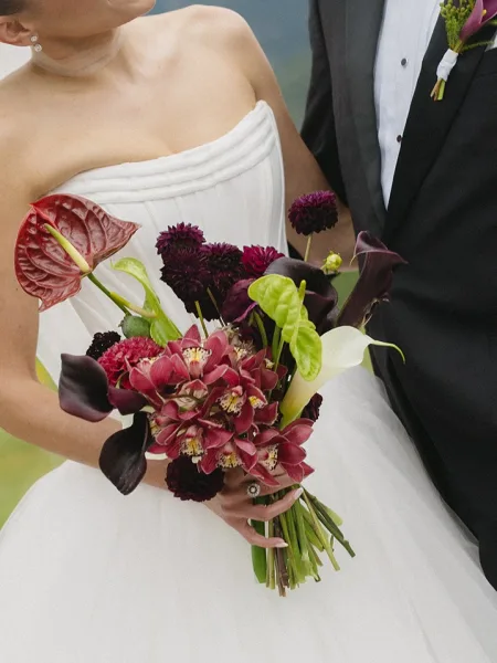 Bridal bouquet, an orchid bridal bouquet with an anthurium accent, held by the bride against outdoor greenery on a hillside.