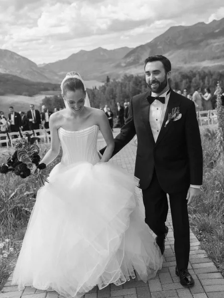 Wedding recessional with the bride and groom walking down a brick path, her bouquet in hand, framed by a mountain range and cloudy sky.