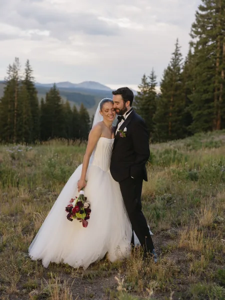 Bride and groom portrait against a mountain backdrop, the couple posed amid pine trees as the bride holds a burgundy bouquet.