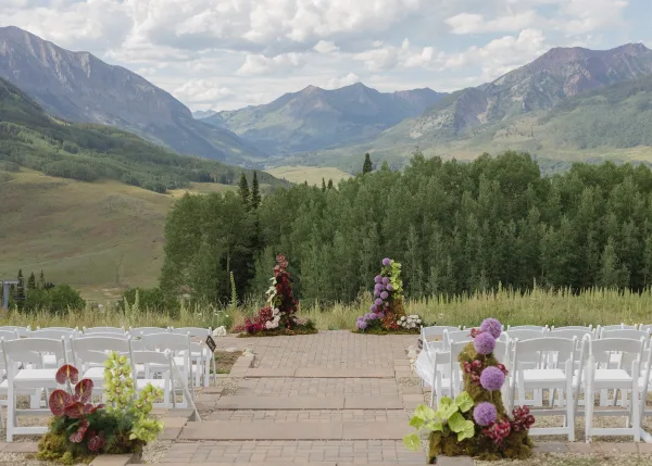 Outdoor ceremony setup for a mountain view ceremony, framed by floral pillars in a meadow with mountains and forest trees beyond.