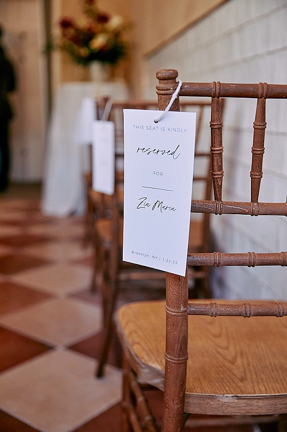 Reserved seat sign tied with string to a wooden Chiavari chair, with ceremony chairs and a floral arrangement on a checkered floor behind