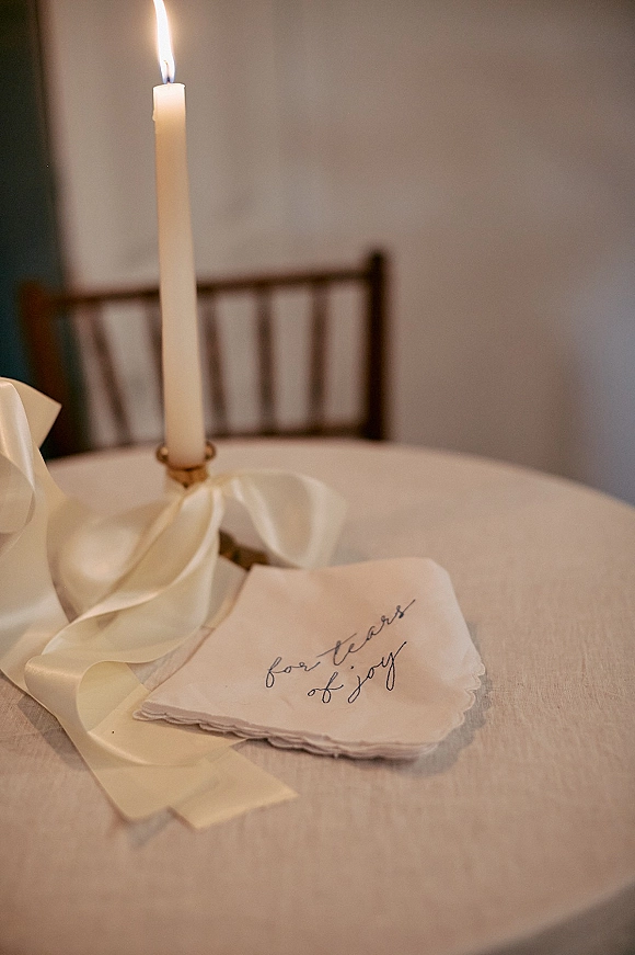 Wedding place setting with a linen napkin featuring calligraphy, tied in silk ribbon beside a taper candle in a holder on a round table indoors