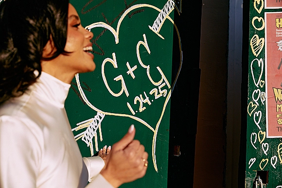 Wedding graffiti wall with wedding initials chalk, heart and arrow drawings, and date text beside a bride’s ringed hand in white dress