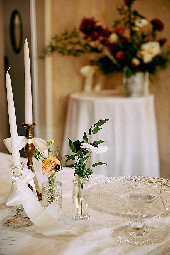 Reception tablescape with wedding bud vase centerpieces, taper candles in brass holders, white ribbons, and crystal cake stand on white linen table